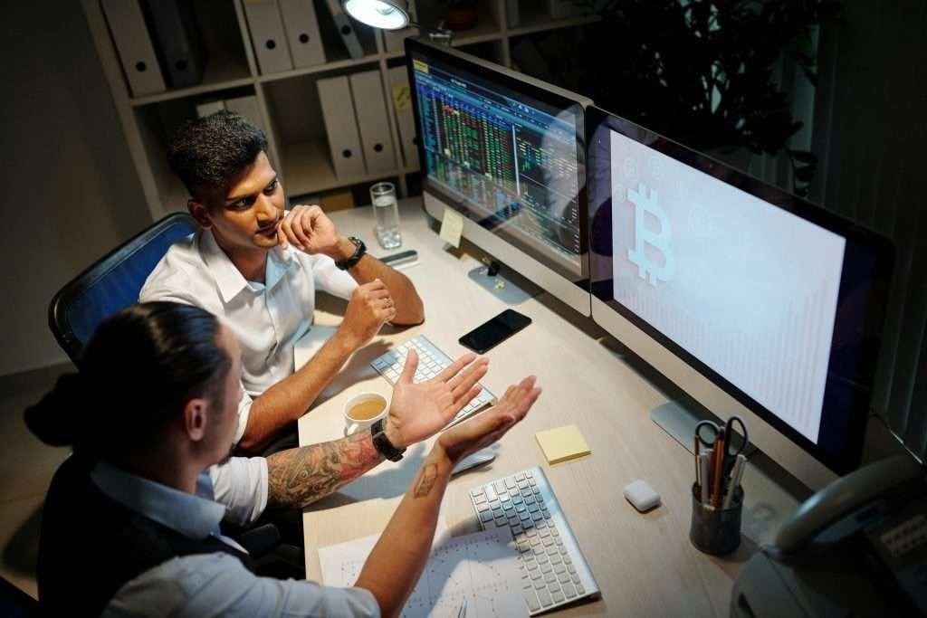 Two male traders at computer screens trading and reviewing brokers